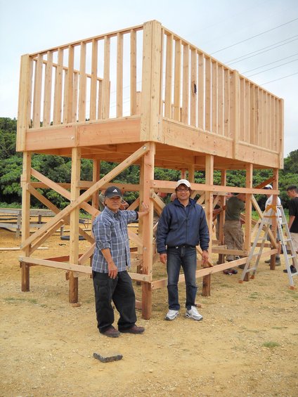 Two men stand next to a wooden structure being built, with others working in the background
