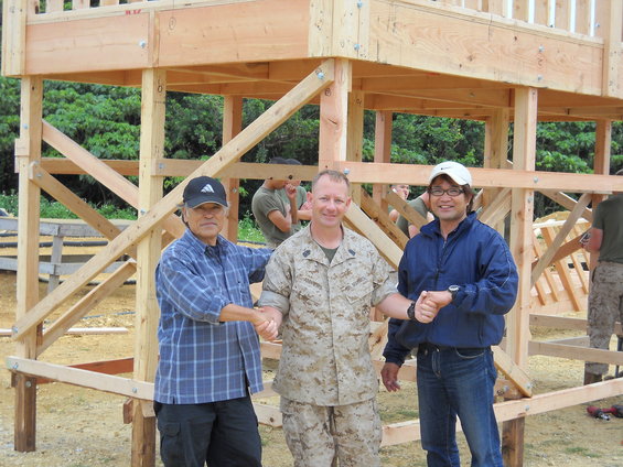 Three men shake hands in front of a wooden structure under construction. One man wears a USMC uniform