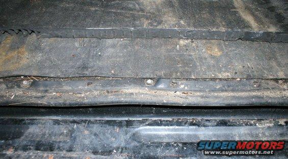 Close-up of the dirty, rusty undercarriage of a vehicle, showing metal panels and some debris