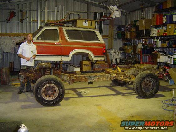 Orange and white Ford Bronco body separated from its rusty chassis in a garage, with a man standing nearby