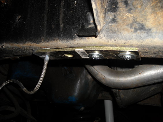 Close-up of a metal plate bolted to a rusty frame rail, with a braided brake line attached. Exhaust pipe visible below