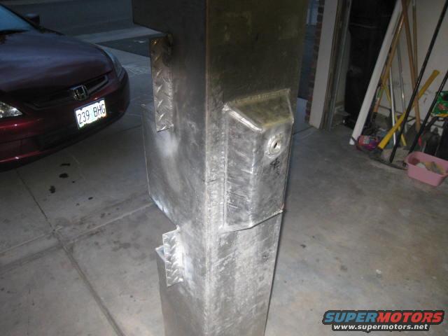 Close-up of a large, rectangular metal fuel tank with visible welds and mounting brackets. A portion of a red car is visible in the background