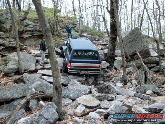 Blue SUV driving over rocks on a wooded trail. Two people are standing nearby