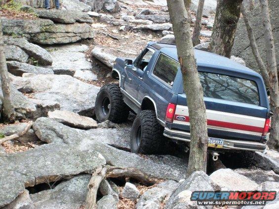 Blue Ford Bronco with large tires navigating rocky terrain, rear view