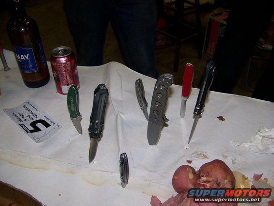 Several folding knives, a beer can, and a bottle on a table with some potatoes