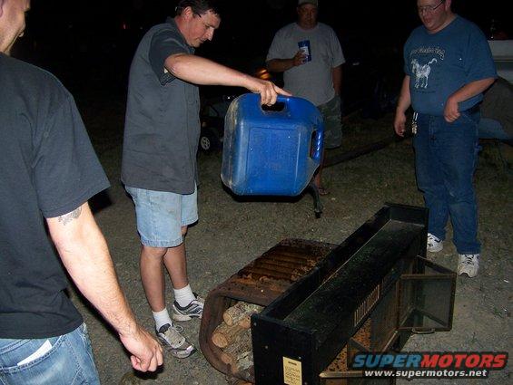 People gathered around a fire pit at night, one person pouring liquid from a blue container
