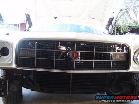 White car's front grille with a red emblem and horizontal bars, showing exposed radiator and tire