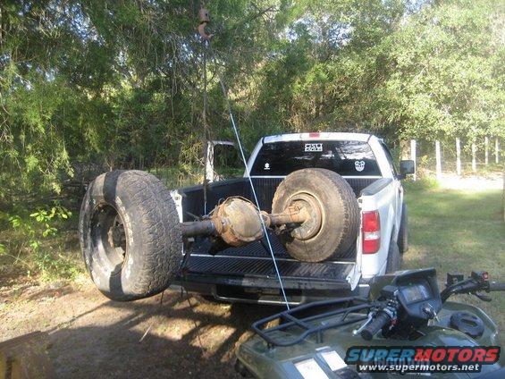 Pickup truck bed holding a vehicle axle with two large tires attached. An ATV is in the foreground