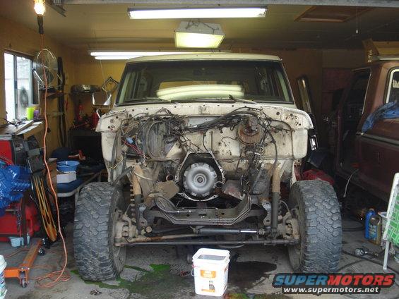 Front view of a white Ford Bronco in a garage, missing its engine and transmission. Another vehicle is parked to the right