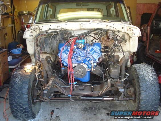 1979 Ford Bronco engine bay with blue engine and front suspension visible. Another vehicle is partially seen on the right