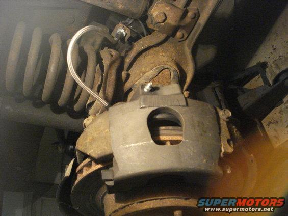 Close-up of a vehicle's front brake caliper and suspension spring. Shows rusty metal components and a silver brake line