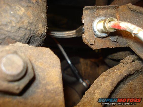 Close-up of a rusty metal bracket with a brake line attached. SuperMotors.net logo visible