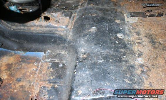 Rusted floor pan inside a vehicle, showing metal corrosion and some wiring