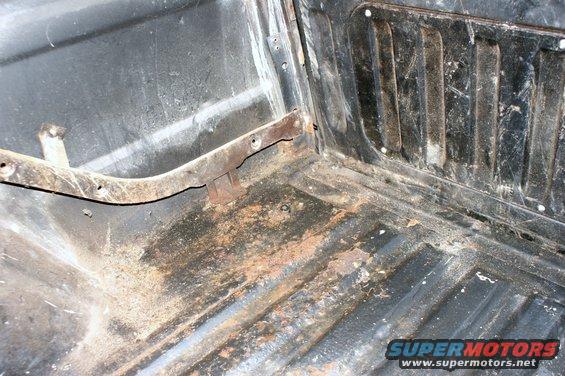 Interior floor of a vehicle, showing rust and dirt. There's a metal bracket attached to the side