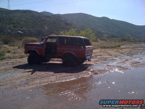 Red 1978 Ford Bronco with door open, driving through mud