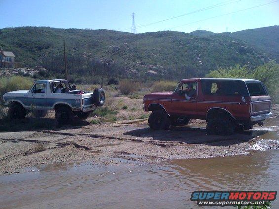 Two lifted 1978 Ford Broncos driving through mud and water, one red, one blue and white