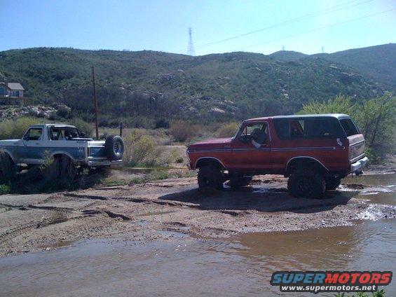Red 1978 Ford Bronco being towed by a blue and white Ford Bronco in a muddy area