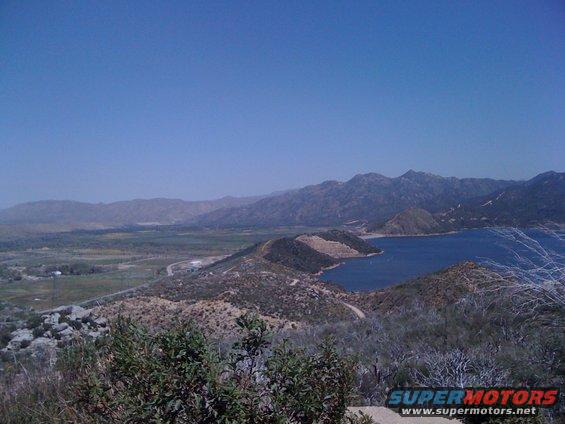 View of a blue lake surrounded by hills and mountains under a clear blue sky. Some vegetation is in the foreground