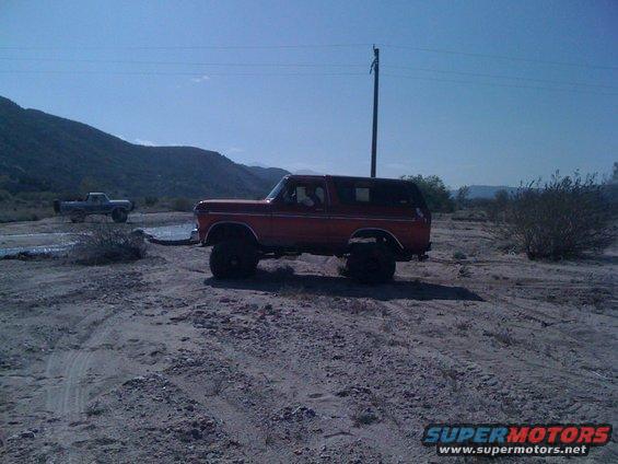 Red 1978 Ford Bronco parked in a dry, dusty area with a utility pole behind it. Another truck is visible in the distance