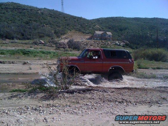 Red 1978 Ford Bronco driving through shallow water, side view, with a house on a hill in the background