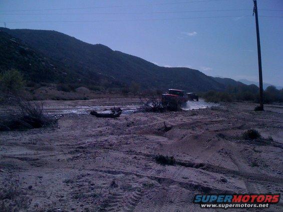 1978 Ford Bronco and another SUV driving through a dusty, dry riverbed with hills in the background