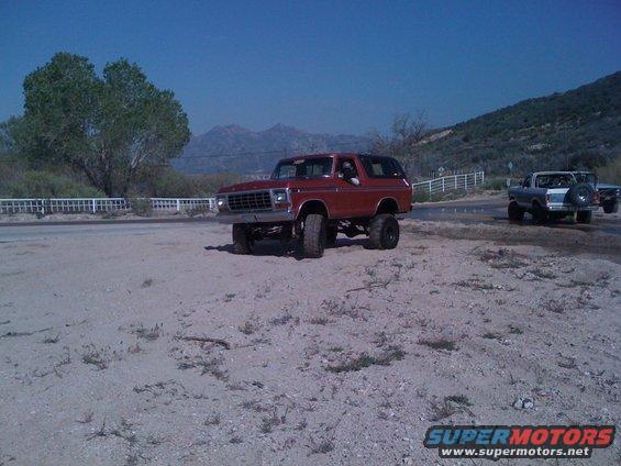 Red 1978 Ford Bronco lifted with big tires, parked on dirt. Another pickup truck is in the background