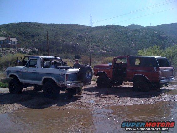 Two Ford Broncos parked in a muddy area with hills in the background. One is blue and white, the other is red