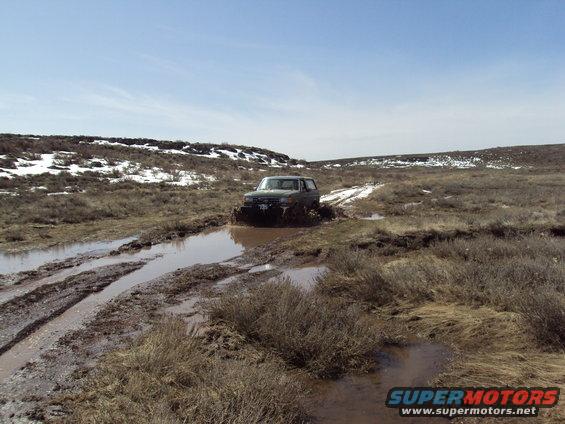 Dark SUV splashing through a muddy track in a barren landscape with snow patches on hills