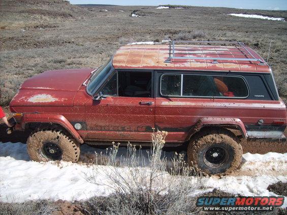 Red Jeep Cherokee Chief stuck in mud and snow, with a roof rack and visible rust on the roof