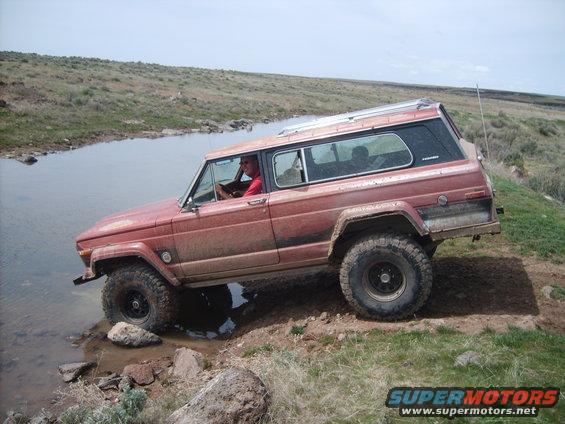 Red Jeep Wagoneer driving through shallow water on a dirt path