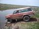 1989 Ford Bronco - A red Jeep Wagoneer navigates a rocky stream bed in a rural, grassy landscape.