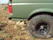 1989 Ford Bronco - The rear of a green SUV is shown on uneven, muddy terrain with rocks. A close-up of the off-road tire is visible.