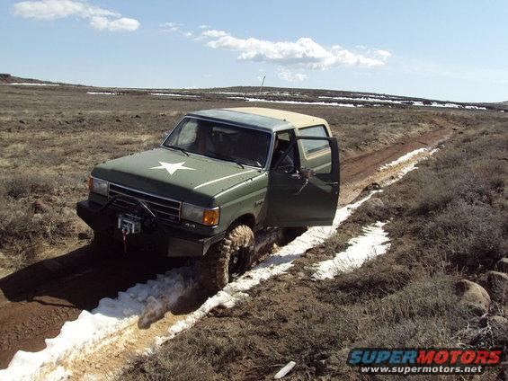 Green Ford Bronco with a white star on the hood, driving off-road with the driver's door open