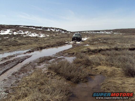 1989 Ford Bronco navigating a muddy track with puddles and sparse vegetation