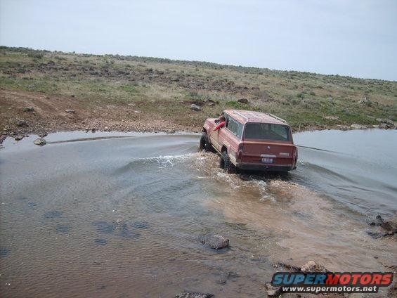 Red Jeep Grand Wagoneer driving through shallow water on a dirt road