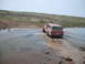 1989 Ford Bronco - A red Jeep Grand Wagoneer navigates through a shallow body of water on a dirt path.