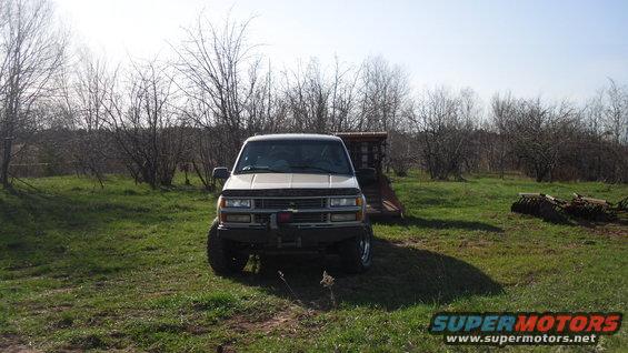 Front view of a lifted beige Chevrolet K1500 pickup truck parked in a grassy field with trees in the background