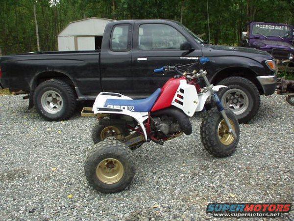 Red, white, and blue 1985 Honda 250 R three-wheeler with gold wheels parked in front of a black pickup truck