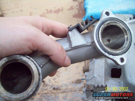 Hand holding a metal intake manifold part with two circular openings and a black plastic clip attached