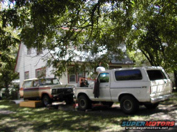 Two Ford Broncos parked in a yard, one white and one tan with red and blue stripes