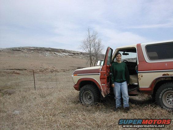 Woman in green sweater and jeans stands by the open driver's door of a red and tan Ford Bronco. Dry grass and a rocky hill are in the background