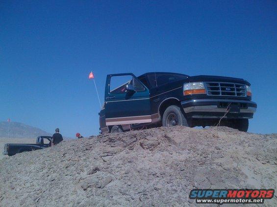 Dark green Ford Bronco with brown trim parked on a dirt hill, driver door open. Another truck is visible in the background