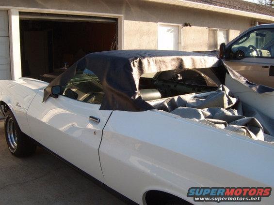 White coupe with a black convertible top cover and a gray car cover draped over the interior. A pickup truck is parked next to it