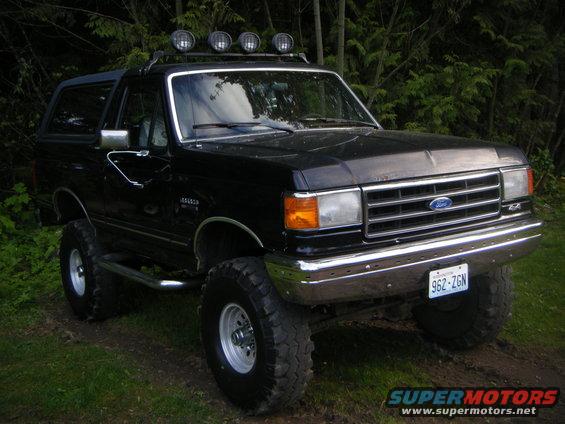 Black Ford Bronco lifted with off-road tires and roof lights, parked on a dirt path with trees in the background