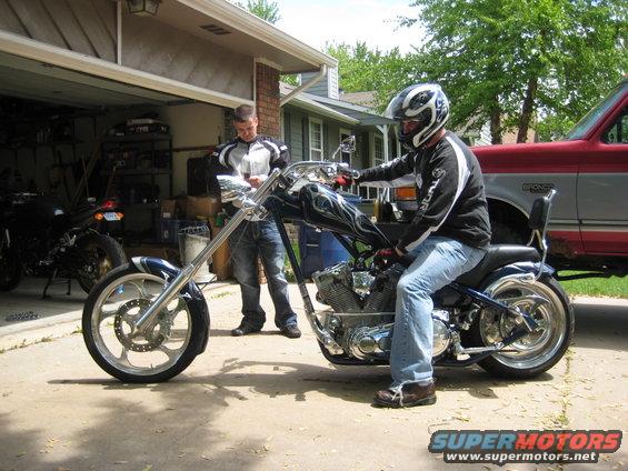 Chopper motorcycle with flame decals on the tank, ridden by a man in jeans and helmet. Another motorcycle is parked in a garage
