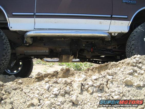Brown and white Ford Bronco side view, showing lifted suspension, side steps, and large tires on dirt