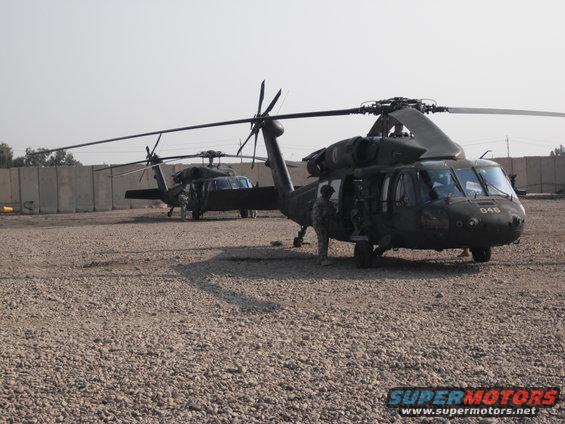 Two military helicopters parked on a gravel lot with concrete barriers in the background