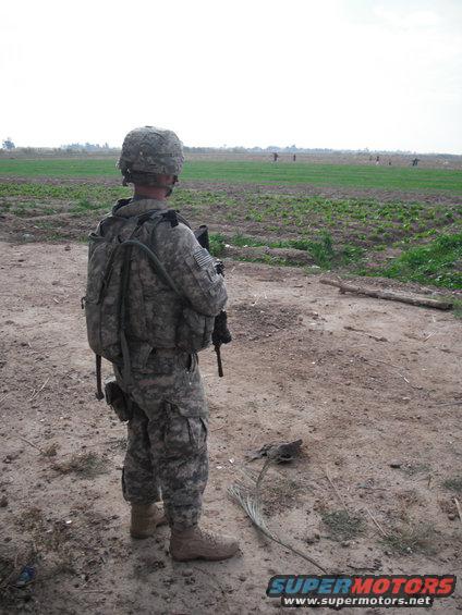 soldier standing in a dirt area looking out at a field with other soldiers in the distance