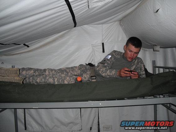 Soldier in camouflage uniform lying on a cot inside a tent, looking at his phone. Another person in uniform is lying down next to him