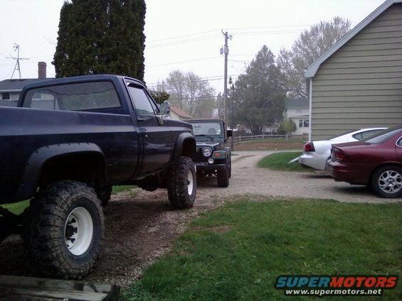 Lifted dark blue pickup truck parked next to a green Jeep. Two cars are parked in the background near a building